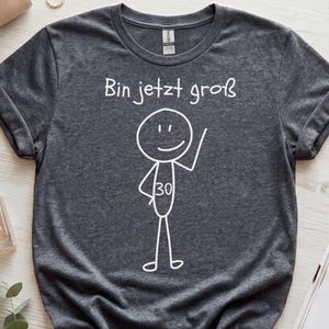 Op de afbeelding: Een donkergrijs gemêleerd T-shirt met korte mouwen, voorzien van een witte afbeelding van een stokfiguurtje met het getal 30 op de romp en de Duitse tekst "Bin jetzt groß" in het wit erboven, met een ronde hals en opgerolde mouwen.