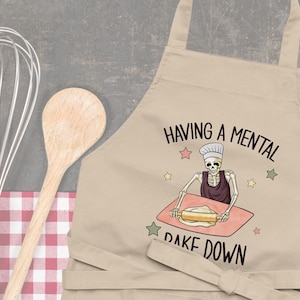 Może przedstawiać: Beżowy fartuch kuchenny z szkieletem piekącym ciasto i tekstem "Having a mental bake down".