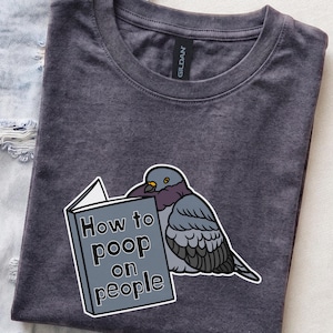 May include: A dark charcoal grey t-shirt featuring a graphic of a pigeon holding a book titled How to poop on people. The bird is illustrated in shades of grey, purple, and black, while the book is a light grey rectangle with black text.