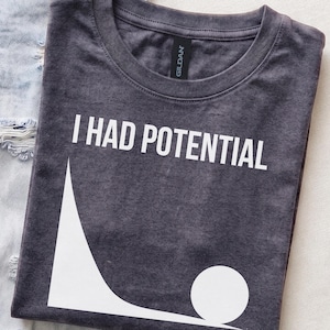 Könnte beinhalten: Ein dunkel anthrazitfarbenes T-Shirt mit Rundhalsausschnitt und dem weißen Schriftzug I HAD POTENTIAL über einer weißen Grafik, die einen Ball am unteren Ende einer geschwungenen Rampe zeigt. Das Gildan-Markenetikett ist am Innenkragen sichtbar.