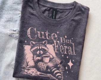 Op de afbeelding: Een antracietgrijs T-shirt met ronde hals en een lichtroze afbeelding van een wasbeer die op een kussen ligt en een mok vasthoudt. De tekst "Cute But Feral" staat in een gestileerd lettertype boven de wasbeer, met kleine sterretjes.