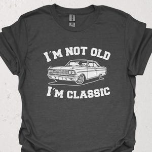 Puede incluir: Camiseta gris oscuro con el texto blanco "I'M NOT OLD I'M CLASSIC". Una ilustración blanca de un coche clásico está centrada debajo del texto. La camiseta es de cuello redondo.