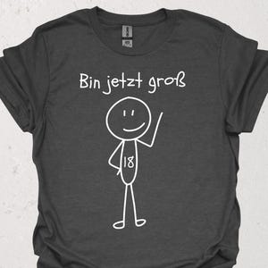 Peut inclure: T-shirt gris foncé avec le texte allemand "Bin jetzt groß" et un bonhomme allumette blanc avec le chiffre 18. Le bonhomme allumette sourit et lève un bras.