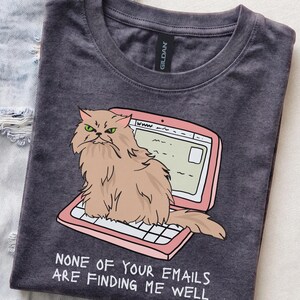 May include: A charcoal gray crew neck t-shirt featuring a graphic of a fluffy tan cat sitting on a pink laptop. The cat has green eyes and a grumpy expression. Below the laptop, the text reads: NONE OF YOUR EMAILS ARE FINDING ME WELL.