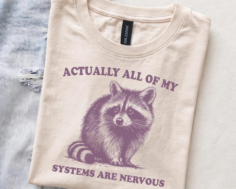 Cremefarbenes Unisex T-Shirt mit einem niedlichen Waschbären und der Aufschrift: Actually All Of My Systems are Nervous.