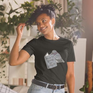 Unisex T-Shirt mit einer Taube die ein Buch liest auf dessen Einband How to poop on people steht.