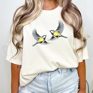 Op de afbeelding: Een crèmekleurig T-shirt met een afbeelding van twee gele en blauwe vogels in de vlucht.