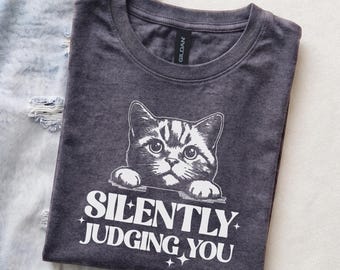 Op de afbeelding: Een donkergrijs T-shirt met ronde hals en een witte afbeelding van een kat die over een rand kijkt, met de tekst "SILENTLY JUDGING YOU" in een vet, gestileerd lettertype eronder, vergezeld van kleine vierpuntige steraccenten.
