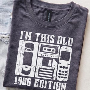 Può includere: Una t-shirt girocollo grigio antracite con una scritta grafica bianca dall'aspetto vissuto che recita I'M THIS OLD 1986 EDITION. Il design include illustrazioni di una cassetta VHS, una musicassetta, un floppy disk e un telefono a conchiglia.