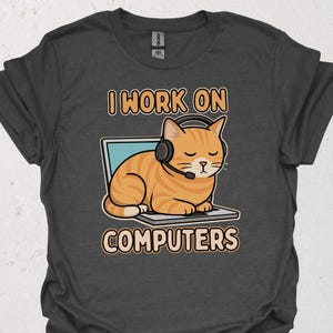 Puede incluir: Camiseta gris oscuro con un gato de dibujos animados que lleva auriculares y está sentado en un portátil. El texto "I WORK ON COMPUTERS" está encima y debajo de la ilustración del gato. El gato es naranja con rayas negras.
