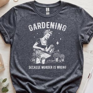 Op de afbeelding: Een donkergrijs t-shirt met korte mouwen en een witte grafische print van een persoon die tuiniert met een gieter. De tekst GARDENING staat boven de illustratie en de tekst BECAUSE MURDER IS WRONG staat eronder.