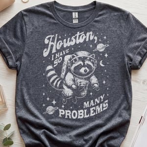 Pode incluir: Uma t-shirt de manga curta em cinzento escuro mesclado com um gráfico branco de um guaxinim com capacete e fato espacial. O texto "Houston, I have so many problems" está rodeado por estrelas, planetas e uma lua crescente num estilo desgastado.