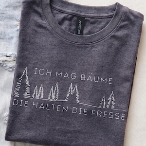 Könnte beinhalten: Ein anthrazitfarbenes T-Shirt mit Rundhalsausschnitt und einer weißen Strichgrafik einer Waldsilhouette. Der Text lautet ICH MAG BÄUME DIE HALTEN DIE FRESSE in einer klaren serifenlosen Schrift. Das Shirt hat ein Gildan-Etikett am Innenkragen.