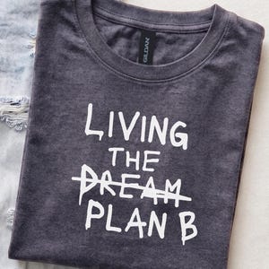 May include: A dark charcoal grey crew neck t-shirt with white screen-printed text that reads LIVING THE DREAM with the word DREAM crossed out by a horizontal line, followed by the text PLAN B underneath in a matching white font style.