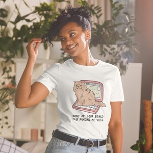 Weißes Unisex T-Shirt mit einer grummeligen Katze die auf einem Laptop sitzt und der Aufschrift: None of Your Emails are Finding me Well.