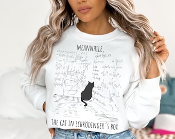 May include: White sweatshirt with a black graphic of a cat and mathematical equations. The text reads "MEANWHILE, THE CAT IN SCHRÖDINGER'S BOX". The long-sleeved top is a casual style.