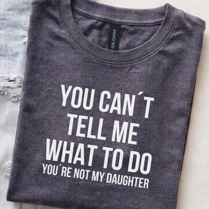 May include: A dark heather grey crew neck t-shirt featuring white text that reads: YOU CAN'T TELL ME WHAT TO DO YOU'RE NOT MY DAUGHTER. The shirt has a black Gildan brand label at the inner neckline and is positioned next to light blue denim fabric.