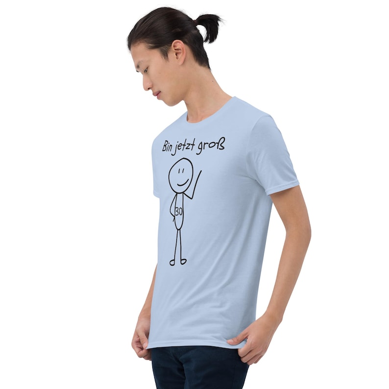 May include: A light blue t-shirt with a stick figure design. The stick figure has a number 30 on its torso and is waving. The text "Bin jetzt gro&szlig;" is above the stick figure. The shirt is being worn by a person.