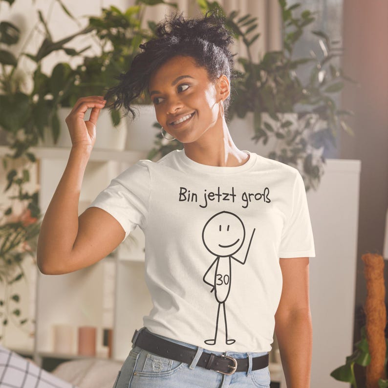 May include: A cream-colored t-shirt with the text "Bin jetzt gro&szlig;" and a stick figure with the number 30. The stick figure is waving. The person is wearing a black belt and blue jeans.