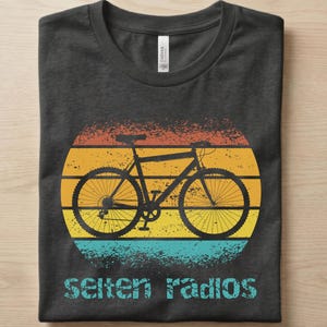 Könnte beinhalten: Ein dunkelgraues T-Shirt mit Rundhalsausschnitt und einer Grafik einer schwarzen Fahrradsilhouette, die in einem abgenutzten, horizontal gestreiften Kreis in Orange-, Gelb- und Türkistönen zentriert ist. Der Text "selten radios" steht in Türkis darunter.