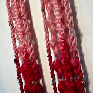 May include: Braided pink and red hair extensions with gold-colored hoops and decorative charms. The hair extensions are a gradient of light pink to deep red. Charms include crystals, moons, and other small beads.