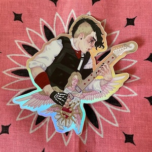 Op de afbeelding: Holografische sticker met een muzikant die gitaar speelt, met een engelenfiguur. De muzikant draagt een zwart vest, rode polsbandjes en een handschoen met skelet. De gitaar heeft hartvormige inlegwerk. De sticker staat op een roze bandana-achtergrond.