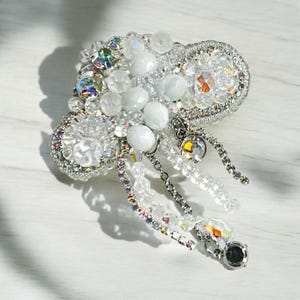 May include: A silver brooch with a white and iridescent crystal design. The brooch is shaped like a cloud with dangling crystal strands.