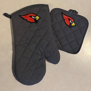 May include: A set of two black quilted oven mitts with a red cardinal logo embroidered on each mitt. The mitts are designed for protecting hands from heat while cooking or baking.