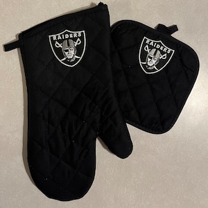 May include: Black oven mitts with a white embroidered Raiders logo. The logo features a football player's helmet and the word "RAIDERS".