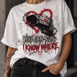 Könnte beinhalten: Ein weißes T-Shirt mit einer Grafik einer Überwachungskamera in einem roten Herz. Der Text lautet: SAME ROUTES SAME PLACES SAME YOU, YOU'RE SAFER WHEN I'M WATCHING und YOU CAN RUN I KNOW WHERE. Das Design enthält rote Tropfen und Stacheldraht.