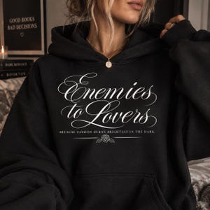 Könnte beinhalten: Ein schwarzer Kapuzenpullover mit weißem Text, der Enemies to Lovers lautet, sowie dem Untertitel BECAUSE PASSION BURNS BRIGHTEST IN THE DARK. Unter dem Text befindet sich eine kleine Strichzeichnung einer Rose über einer Linie.