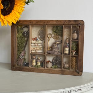 May include: A wooden display box with various compartments, containing miniature witch-themed items. Features a broom, books, bottles, a key, a hat, and a spider. The box is brown and rests on a white surface. A sunflower is in the background.