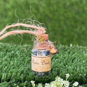 May include: A small clear glass bottle filled with iridescent glitter, topped with a cork stopper and a small metal bell. A tan label with the text Pixie Dust is wrapped around the centre. A frayed peach-coloured twine is tied around the neck.
