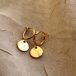 Peut inclure: Une paire de boucles d'oreilles créoles dorées avec une texture martelée. Chaque boucle d'oreille est ornée d'un petit charme rond. Les boucles d'oreilles sont présentées sur un fond en tissu beige.