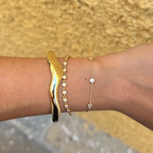 Könnte beinhalten: Ein goldfarbenes Armband-Set. Das Set enthält ein breites, wellenförmiges Armband, ein zartes Kettenarmband mit kleinen, runden Verzierungen und ein dünnes Kettenarmband mit kleinen, runden, klaren Steinen. Die Armbänder werden an einem Handgelenk getragen.