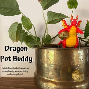 May include: A red and yellow dragon figurine holding a gold 20-sided die, perched on the edge of a brass pot with a green plant. The pot has a gold 20-sided die hanging from it. The text "Dragon Pot Buddy" is written in black on a white background. The text "Painted version is shown as an example only. Your pot buddy arrives unpainted." is written in black on a white background.