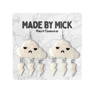 Może przedstawiać: Białe kolczyki w kształcie chmury ze smutną buźką i trzema zawieszkami w kształcie błyskawicy wiszącymi z każdej chmury. Kolczyki znajdują się na białej kartce z tekstem "MADE BY MICK @mick_themaker".