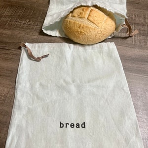 May include: Two white cotton bread bags with the word "bread" printed on them. One bag is empty and the other contains a loaf of bread.