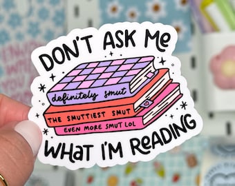 May include: A white sticker with the text "DON'T ASK ME WHAT I'M READING" and a stack of books. The books are pink, red, and purple with the words "definitely smut", "THE SMUTTIEST SMUT", and "EVEN MORE SMUT LOL".