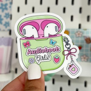 Puede incluir: Una pegatina con un diseño verde y rosa de una funda de auriculares inalámbricos con el texto "Audiobook Girlie". La pegatina incluye un lazo, una mariposa y una fresa. Los auriculares son blancos.