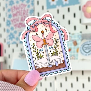 May include: A whimsical sticker featuring a sword piercing a book, with a butterfly design and a pink bow. The sticker has a light blue scalloped border and includes stars and floral accents. The overall design is in soft pastel colors.
