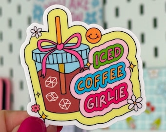 May include: A colorful sticker featuring an iced coffee drink with a straw and a pink bow. The text on the sticker reads "Iced Coffee Girlie" in bubble letters. The sticker has a yellow border and small flower and star accents.
