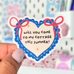 Puede incluir: Una pegatina en forma de corazón con un borde festoneado azul y lazos rojos. La pegatina tiene el texto "Will you come to my cottage this summer?" en negro. El fondo es de color crema claro con dos pequeñas estrellas doradas.