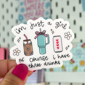 Puede incluir: Una pegatina blanca con el texto "I'm just a girl of course I have three drinks". La pegatina presenta ilustraciones de una bebida en un tarro, un vaso y una lata con corazones. La pegatina está sujeta por una mano con esmalte de uñas rosa.