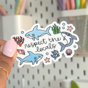 May include: A white sticker with the text "respect the locals" and illustrations of sharks, coral, seaweed, and starfish. The sharks are light blue and the other elements are in various colors.