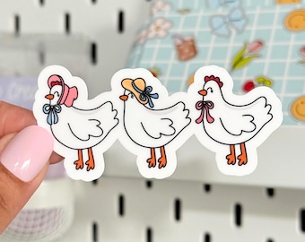 May include: Three white chicken stickers with cartoon-style illustrations. Each chicken wears a different hat: a pink bonnet, a yellow sun hat, and a red comb. The stickers have a white border and are held by a hand.