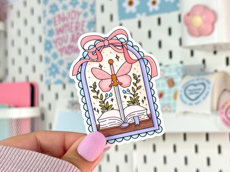 May include: A sticker featuring a whimsical design with a sword piercing an open book. A pink butterfly with a sword body is centered above the book. The sticker is framed with a light blue scalloped border and a pink bow at the top. Green leaves and stars complete the design.