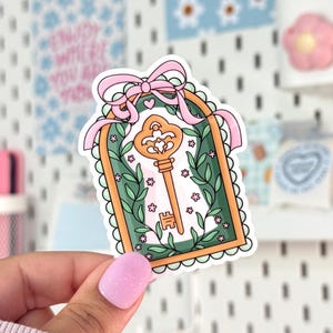 May include: A sticker featuring a gold key inside a green and orange frame, adorned with pink flowers and green vines. A pink ribbon with a heart detail tops the frame. The sticker has a scalloped edge.