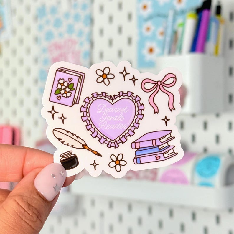 May include: A sticker featuring a heart with the text "Dearest Gentle Reader", books, flowers, a quill, and a bow. The sticker has a white background with a variety of pastel colors, including pink, purple, and black. The sticker is held by a person.
