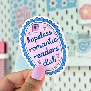Peut inclure: Autocollant ovale rose avec une bordure bleue festonn&eacute;e. Il affiche l'inscription &laquo; hopeless romantic readers club &raquo; dans une police manuscrite, avec des illustrations de c&oelig;urs et de livres.